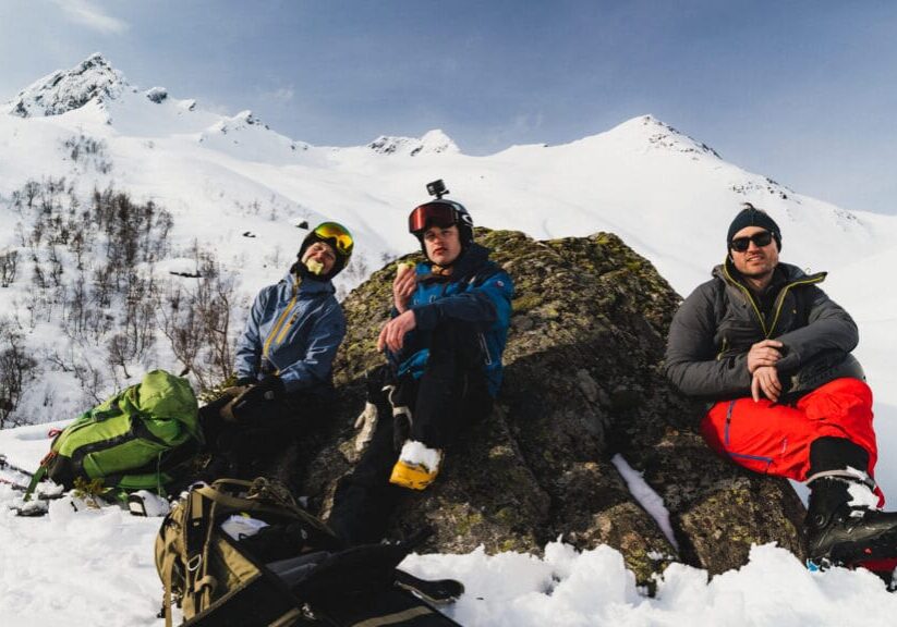 Photo by Alex Baker Guiden Tabitha med to venner har matpause mitt i vinterfjellet med sol i fleisen.